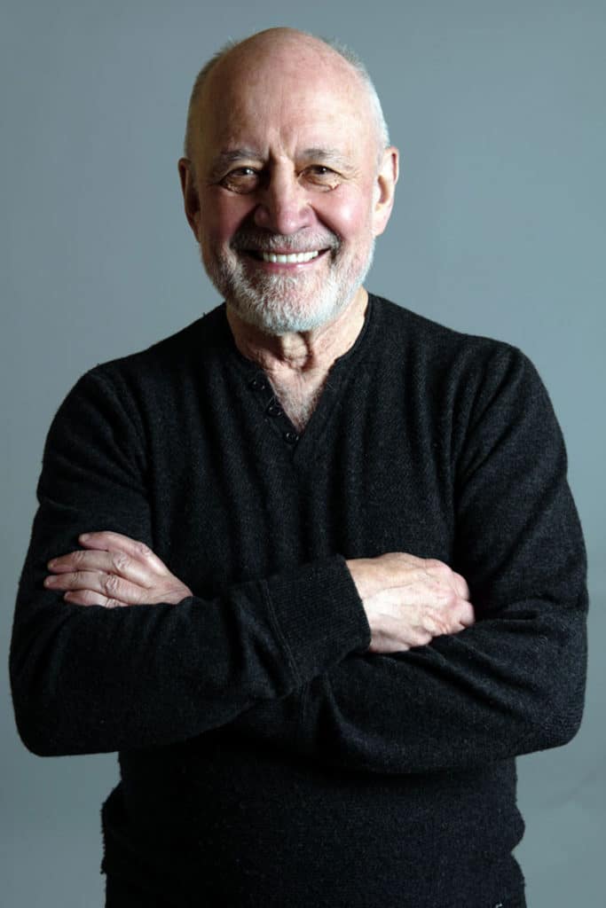 headshot of older man folding arms smiling widely with dark grey background